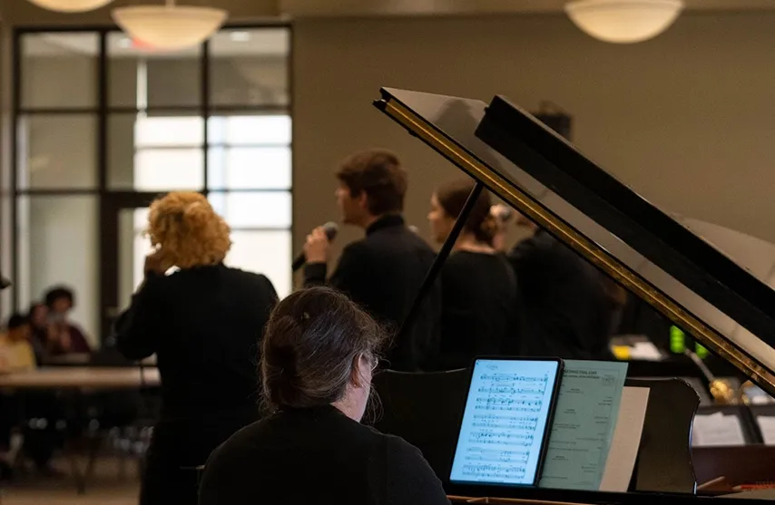 A pianist plays from sheet music in the foreground while SAU students sign into microphones in the background.