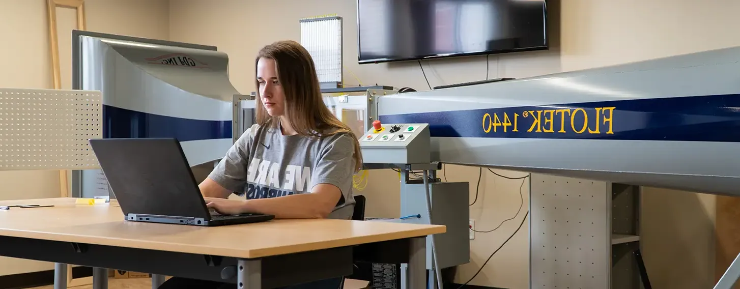 A female engineer sitting at a computer with a large wind tunnel in the background.